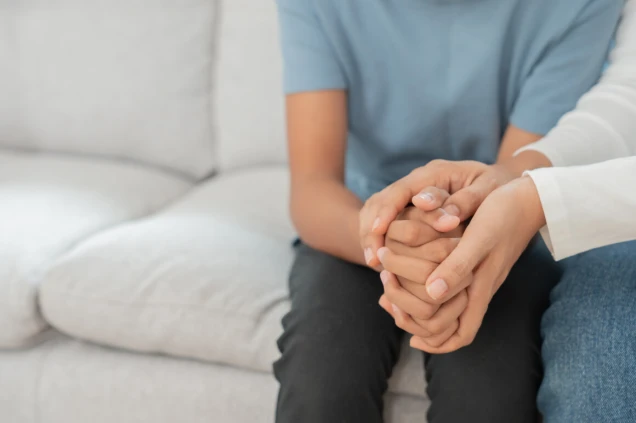 Zwei Personen sitzen auf einem Sofa. Eine Person in einem blauen Oberteil hält die Hände der anderen Person, die ein weißes Oberteil trägt.