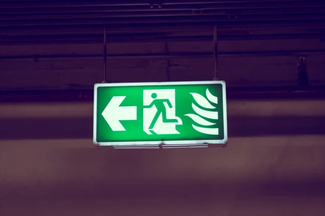 Grünes beleuchtetes Fluchtweg-Schild mit einer Person, die durch eine Tür nach links läuft, und Flammen-Symbol auf der rechten Seite, hängend an der Decke.
