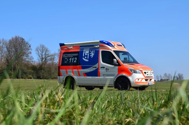 Ein Rettungswagen mit orange-weißer Lackierung und blauen Akzenten steht auf einer Wiese. Im Hintergrund sind Bäume und ein klarer blauer Himmel sichtbar.