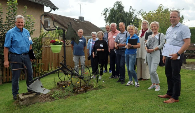 Eine Gruppe von Personen steht auf einer Wiese neben einem altes landwirtschaftliches Gerät, das mit Blumen dekoriert ist. Im Hintergrund sind Häuser und Bäume zu sehen.