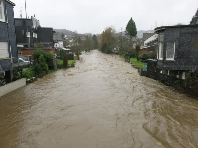 Überflutete Agger, die durch eine Wohngegend mit Häusern auf beiden Seiten fließt. Das Wasser reicht bis zu den angrenzenden Grundstücken. Trübes Wetter.