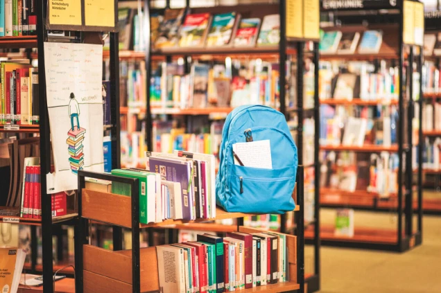 Ein blauer Rucksack steht auf einem Bücherregal in einer Bibliothek. Im Hintergrund sind weitere Regale mit Büchern zu sehen, teils unscharf. Ein Poster mit einer Zeichnung einer Person auf einem Bücherstapel hängt an einem Regal.