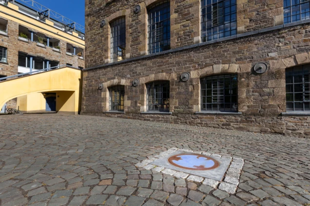 Ein gepflasterter Hof mit einem runden Kunstwerk im Boden, umgeben von einem quadratischen Rahmen. Links ein gelbes Gebäude, rechts ein Backsteinbau mit großen Fenstern.