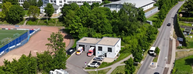 Luftaufnahme eines Gebäudes mit weißen Wänden und Flachdach, davor geparkte Rettungswagen. Rechts eine Straße mit Autos, links ein Sportplatz mit Kunstrasen.