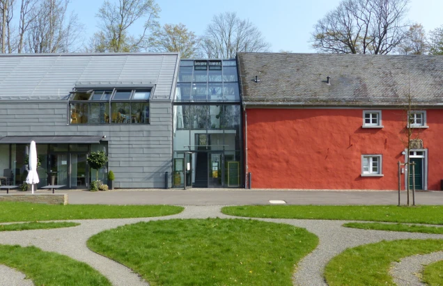 Modernisiertes Gebäude mit einer Kombination aus grauem Metall und Glas sowie einem roten Anbau. Vor dem Gebäude befinden sich ein Weg und grüne Rasenflächen.