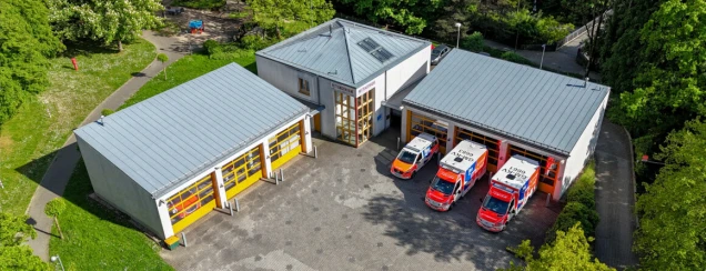 Luftaufnahme einer Rettungswache mit zwei Gebäuden. Vor einer Garage stehen drei Einsatzfahrzeuge. Umgeben von Grünflächen und Bäumen.
