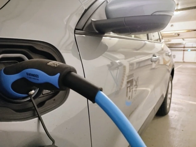 Ein Elektroauto wird in einer Tiefgarage mit einem blauen Ladekabel aufgeladen. Der Ladeanschluss und ein Seitenspiegel des Autos sind sichtbar.