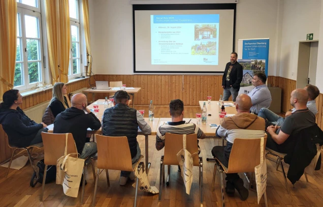 Ein Seminarraum mit einer Gruppe von Personen, die an Tischen sitzen und einem Vortrag zuhören. Im Hintergrund eine Präsentationsfolie und ein Redner.