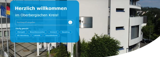 Willkommensbildschirm mit der Aufschrift 'Herzlich willkommen im Oberbergischen Kreis!' in weißer Schrift auf blauem Hintergrund. Darunter ein Suchfeld mit der Eingabeaufforderung 'Suchbegriff eingeben' und häufig gesuchten Themen wie Elterngeld, Wunschkennzeichen und Gesundheitsamt. Im Hintergrund ein modernes, weißes Gebäude mit blauen Fenstern, davor Flaggenmasten mit wehenden Flaggen sowie Büsche und ein Parkplatz.