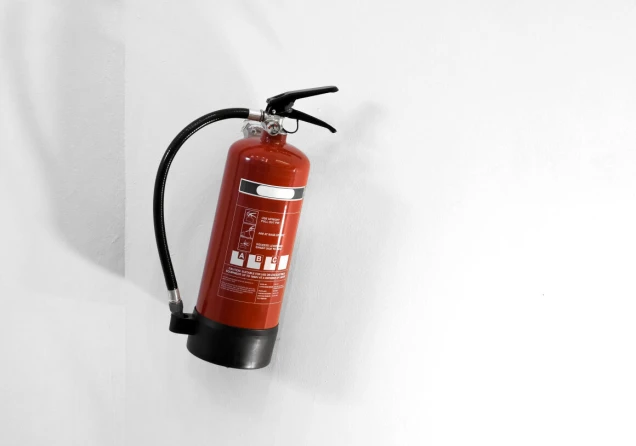 Ein roter Feuerlöscher mit schwarzem Schlauch und Hebel, montiert an einer weißen Wand.