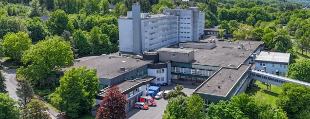 Luftaufnahme eines mehrstöckigen Gebäudekomplexes, umgeben von vielen Bäumen. Vor dem Gebäude stehen mehrere Einsatzfahrzeuge.