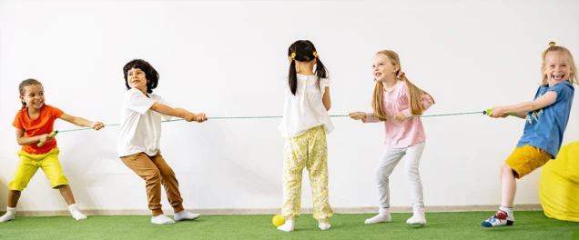 Kinder spielen Tauziehen auf grünem Boden in einem hellen Raum. Zwei Teams ziehen an einem Seil, jedes Team besteht aus zwei Kindern, ein Kind steht in der Mitte.