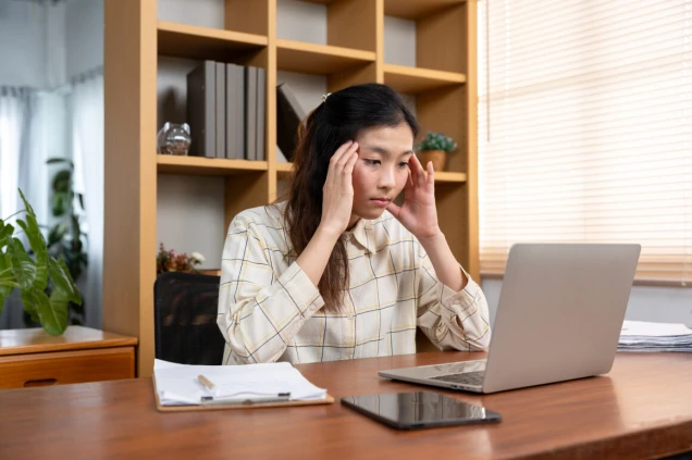 Stressed young Asian woman touching her temples while looking at laptop screen during remote work, overwhelmed with workload and digital pressure in a home office setup.