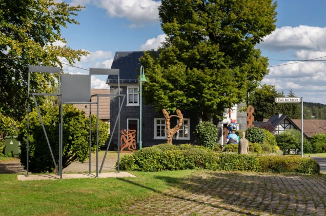Kleiner Platz mit verschiedenen Kunstskulpturen, darunter eine abstrakte Metallstruktur, eine Holzskulptur und eine bunte Figur. Im Hintergrund ein Gebäude und Bäume.