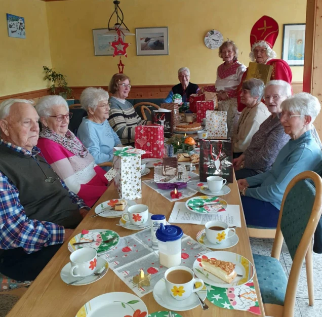 Eine Gruppe älterer Menschen sitzt an einem festlich gedeckten Tisch mit weihnachtlicher Dekoration, Geschenken und Kaffee. Zwei Personen in Nikolauskostümen stehen dahinter.