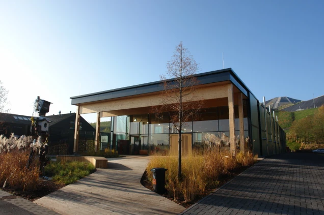 Modernes Gebäude mit Holz- und Glasfassade, umgeben von Pflanzen und einer Holzplattform.