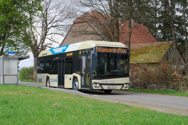 Ein Wasserstoffbus der OVAG steht an einer Haltestelle auf einer Landstraße. Im Hintergrund sind Bäume und ein Gebäude mit Ziegeldach zu sehen.