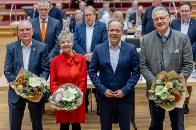 Vier Personen stehen in einer Reihe in einem Saal. Zwei halten Blumensträuße, eine trägt ein rotes Kleid. Im Hintergrund sitzen weitere Personen an Tischen.
