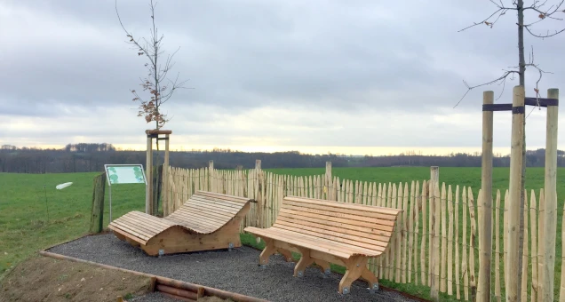 Holzliege und Holzbank auf einem kleinen mit Kies belegten Bereich, umgeben von einem niedrigen Holzzaun. Zwei junge Bäume mit Stützpfählen. Im Hintergrund eine Wiese und bewölkter Himmel.