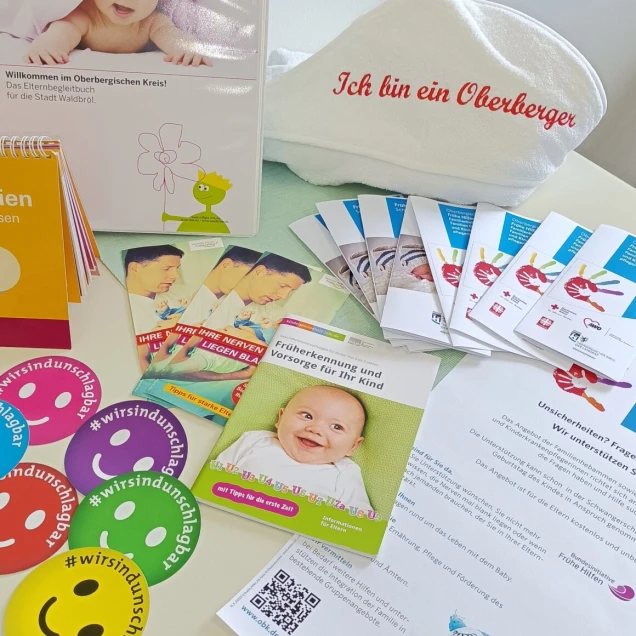 Verschiedene Informationsmaterialien und Flyer rund um Familie und Kinderbetreuung, darunter ein Handtuch mit der Aufschrift 'Ich bin ein Oberberger' und bunte Sticker.