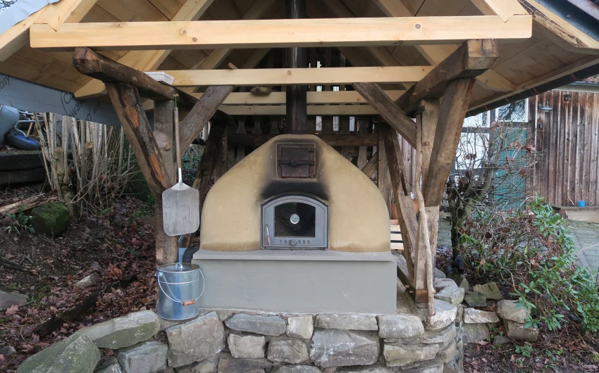 Ein mit Holz überdachter Lehmofen mit Metalltür und Schornstein. Daneben ein Pizzaschieber und ein Eimer. Der Ofen steht auf einem gemauerten Steinsockel.