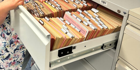 Eine Person in einem gemusterten Hemd steht vor einem offenen Aktenschrank und sortiert oder betrachtet Akten in einer Schublade mit farbigen Hängeregistern.