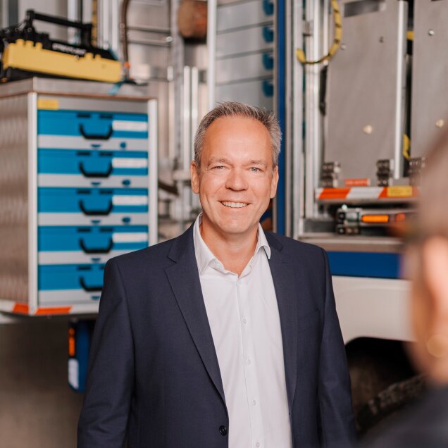 Ein Mann in einem Anzug steht lächelnd in einer industriellen Umgebung mit Regalsystemen und technischen Geräten im Hintergrund.