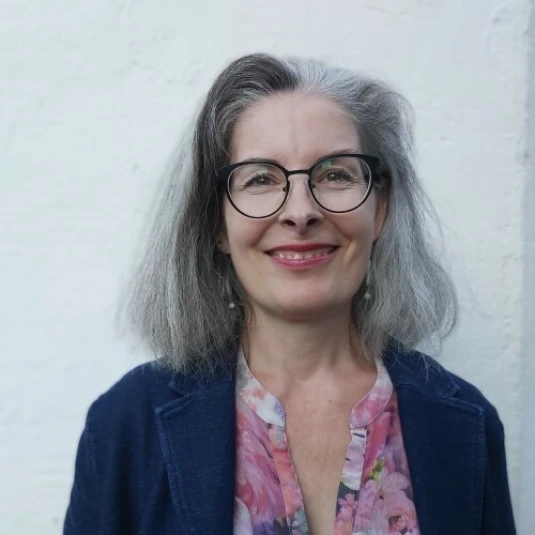 Eine Frau mit grauem Haar, Brille und einem freundlichen Lächeln steht vor einem weißen Hintergrund. Sie trägt eine blaue Jacke und eine bunte Bluse.