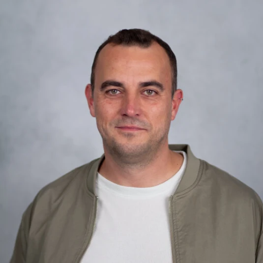 Porträt eines Mannes mit kurzem dunklem Haar, der ein weißes T-Shirt und eine olivgrüne Jacke trägt. Der Hintergrund ist einfarbig grau.