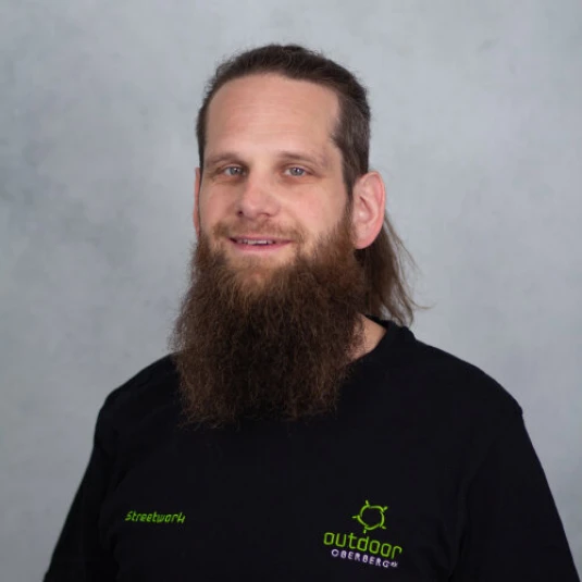 Ein Mann mit langen Haaren und einem langen, vollen Bart trägt ein schwarzes T-Shirt mit grüner und weißer Aufschrift. Der Hintergrund ist grau.