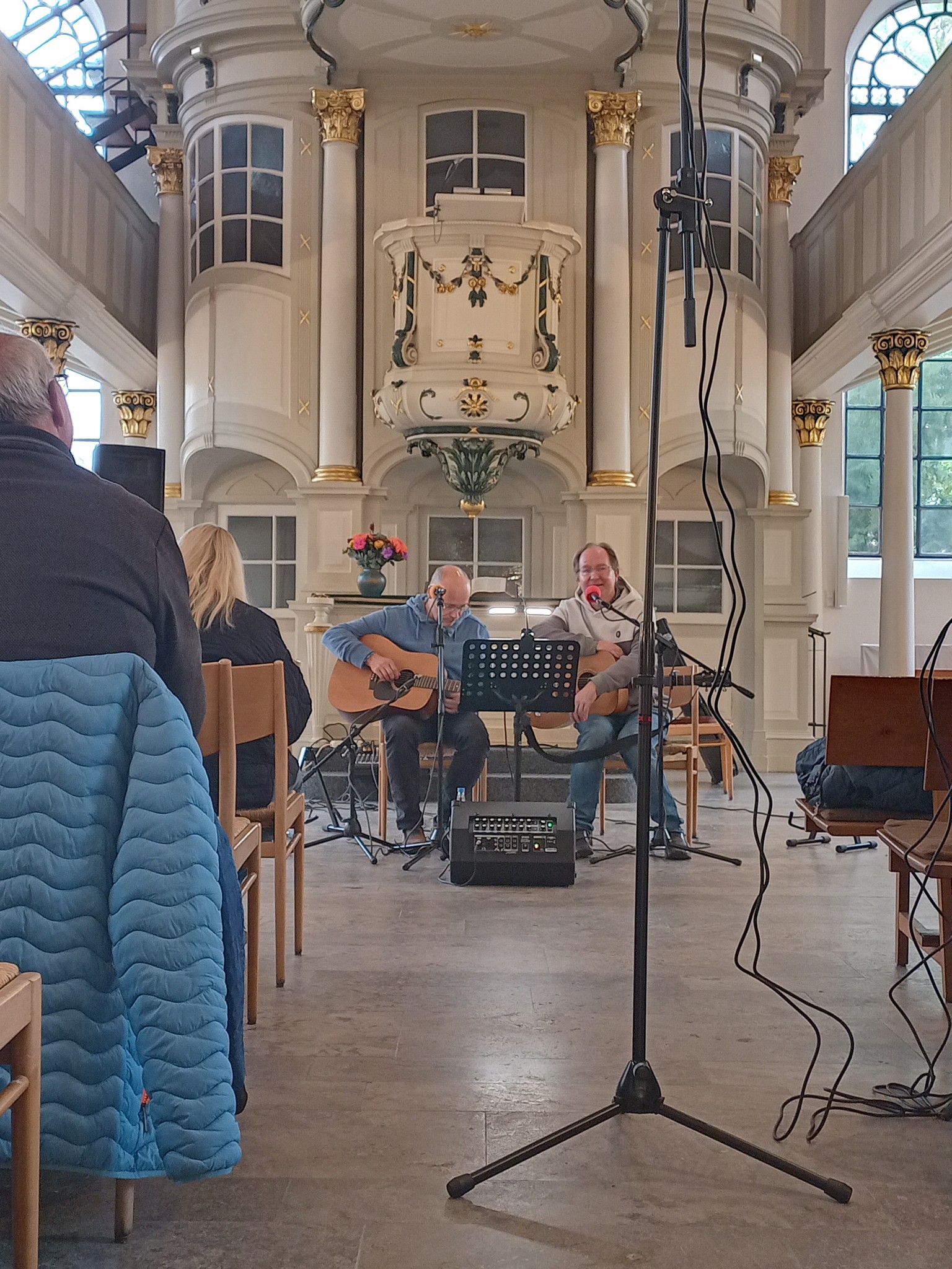 Innenraum einer Kirche mit zwei Musikern, die auf Stühlen vor einem Altar sitzen und Gitarre spielen. Mikrofone und Lautsprecher sind sichtbar, Publikum sitzt auf Holzstühlen.