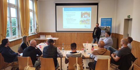 Ein Seminarraum mit mehreren Personen, die um einen Tisch sitzen und einem Vortragenden zuhören. Im Hintergrund eine Präsentation auf einer Leinwand.