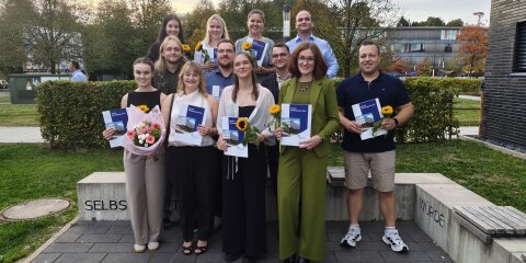Eine Gruppe von zwölf Personen steht lächelnd in einer Außenumgebung vor einer Hecke und Bäumen. Einige halten Blumen und Zertifikate in den Händen.