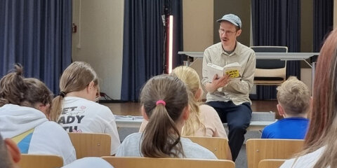 Ein Mann mit Mütze sitzt vor einem Publikum aus Kindern und liest aus einem Buch vor. Der Raum hat blaue Vorhänge und einen Tisch im Hintergrund.