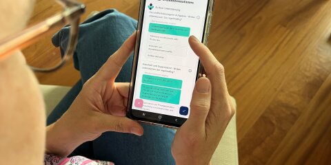 Eine Person hält ein Smartphone in der Hand und betrachtet eine App mit Formularfeldern. Die Person trägt eine Brille und ein gemustertes Hemd. Im Hintergrund ist ein Holzboden sichtbar.