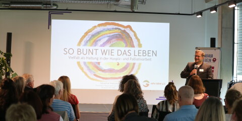 Ein Mann hält eine Präsentation vor Publikum in einem Konferenzraum. Auf der Leinwand steht 'So bunt wie das Leben - Vielfalt als Haltung in der Hospiz- und Palliativarbeit'.