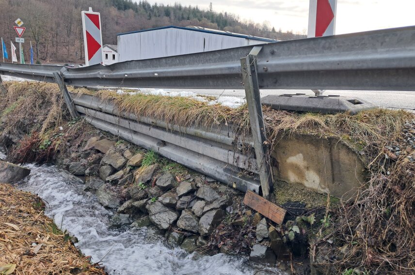 Eine beschädigte Leitplanke an einer Straße neben einem kleinen Bach. Die Böschung ist mit Steinen befestigt, einige Pflanzen wachsen am Ufer. Im Hintergrund sind ein Gebäude und ein bewaldeter Hügel zu sehen.