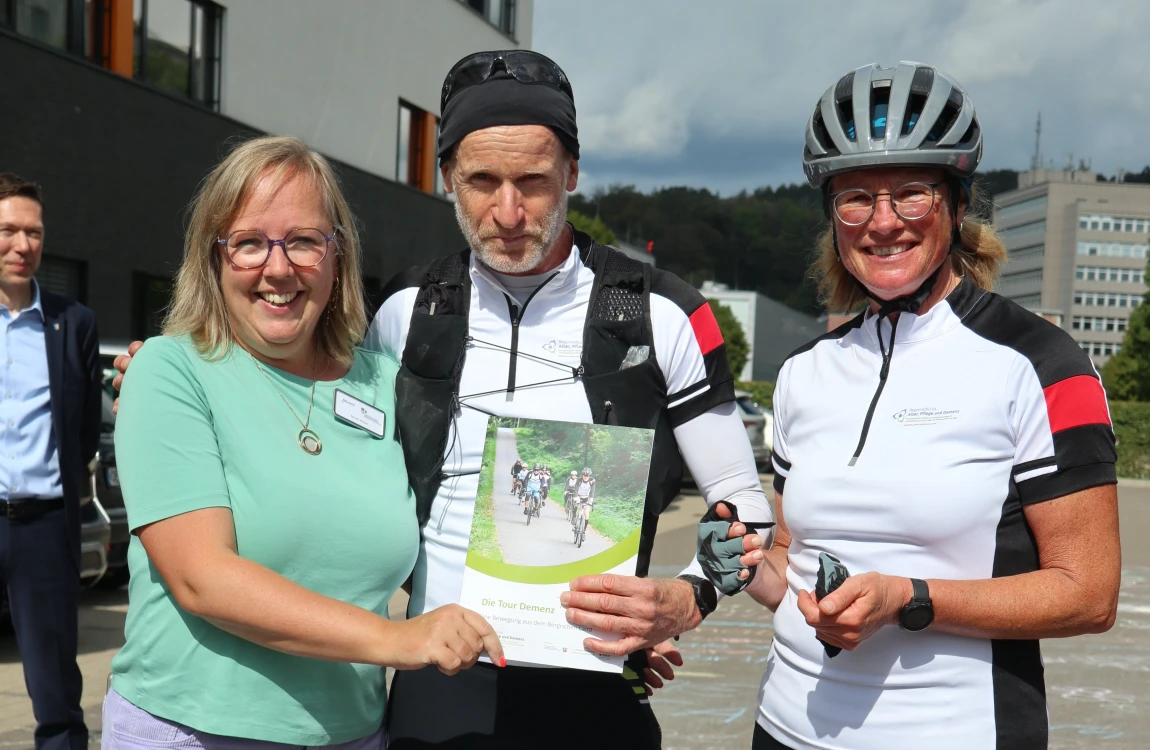 Drei Personen stehen draußen vor einem Gebäude. Zwei tragen Radtrikots, Helme und sportliche Kleidung, eine Person hält ein Blatt Papier mit einem Foto darauf. Die dritte Person trägt Alltagskleidung und lächelt. Im Hintergrund sind weitere Personen, ein Parkplatz und ein Gebäude sichtbar.