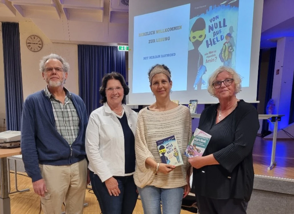 Vier Personen stehen nebeneinander in einem Raum mit Bühne. Zwei Frauen in der Mitte halten Bücher. Im Hintergrund eine Leinwand mit Buchcover.