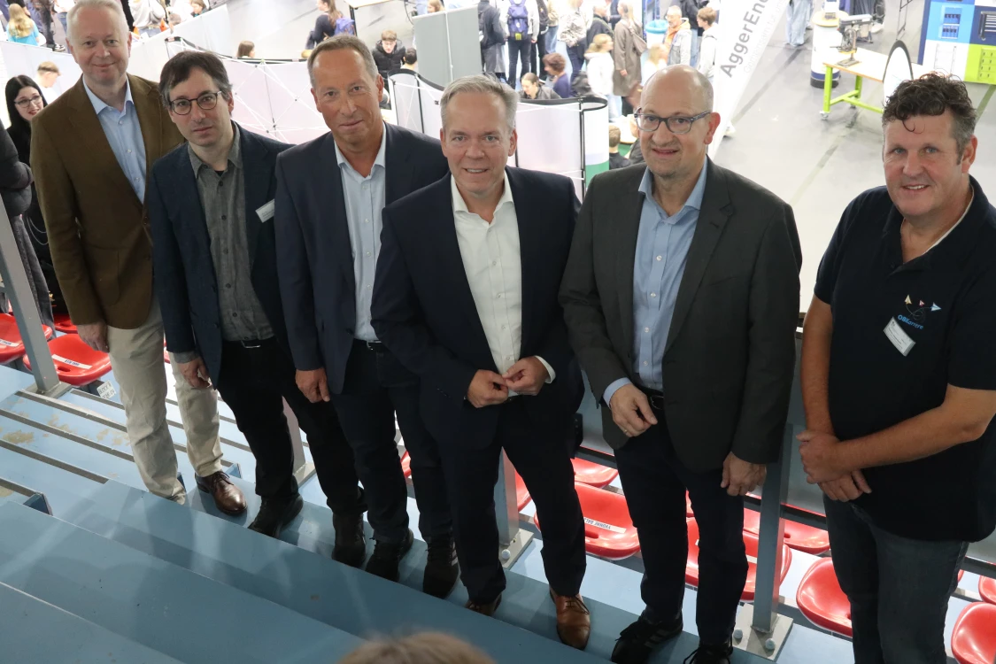 Sechs Männer stehen in einer Reihe auf einer Treppe in einem Innenraum. Hinter ihnen sind Menschen und Messestände zu sehen.