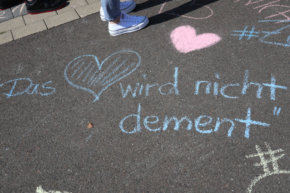 Straßenoberfläche mit Kreidezeichnung. Ein blaues Herz und der Text 'Das Herz wird nicht dement' sind zu sehen. Im Hintergrund Schuhe von Personen.