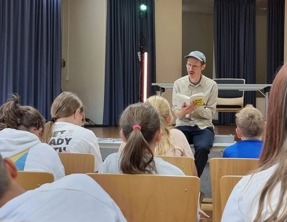 Ein Mann mit Mütze sitzt vor einem Publikum aus Kindern und liest aus einem Buch vor. Der Raum hat blaue Vorhänge und einen Tisch im Hintergrund.