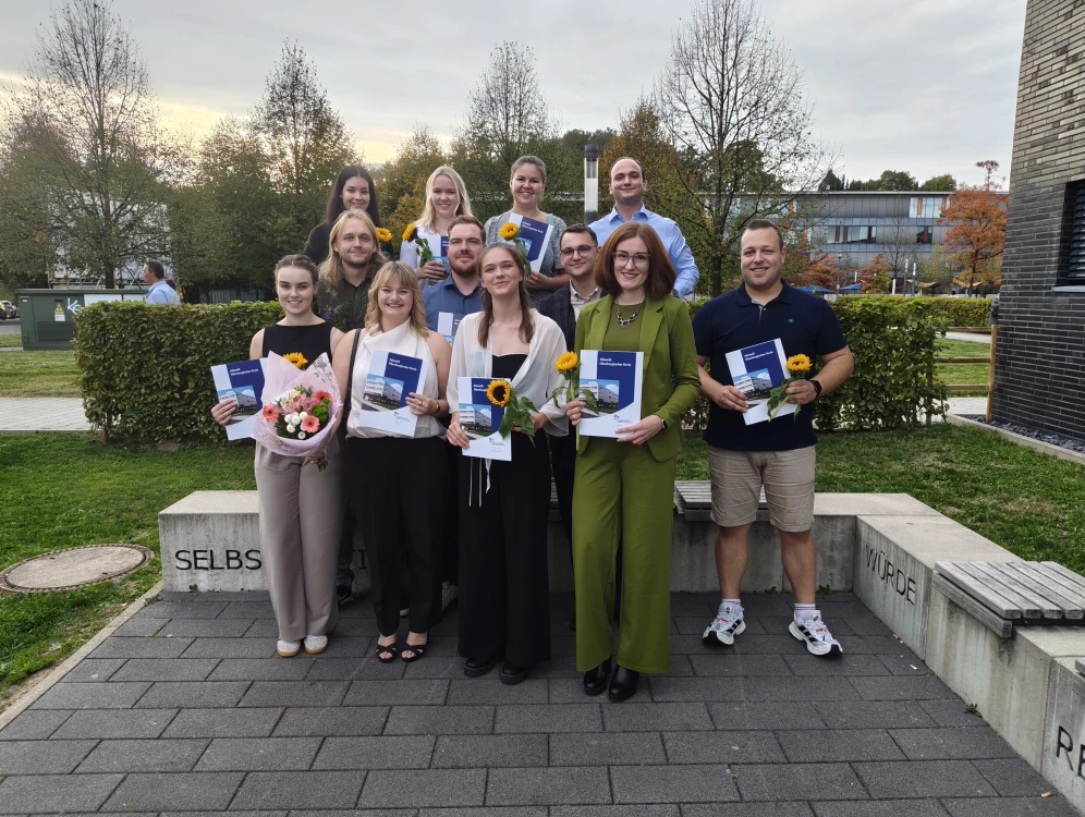 Eine Gruppe von zwölf Personen steht lächelnd in einer Außenumgebung vor einer Hecke und Bäumen. Einige halten Blumen und Zertifikate in den Händen.