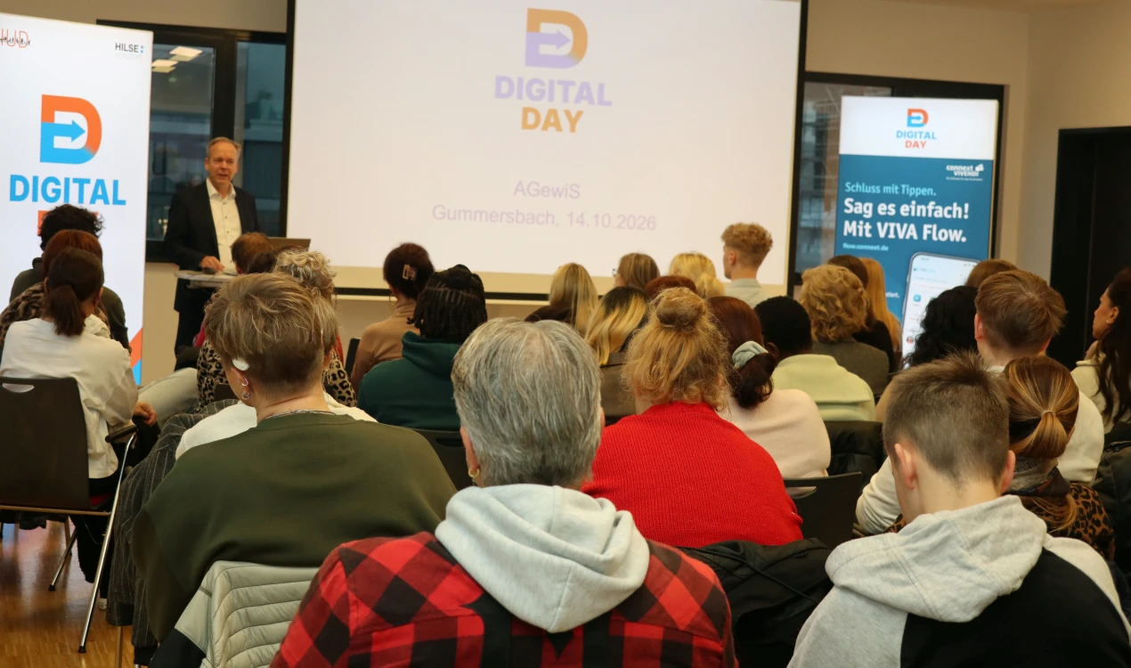 Ein Mann steht an einem Rednerpult vor einer Leinwand, auf der 'Digital Day' und ein Datum angezeigt werden. Publikum sitzt in Reihen und hört zu.