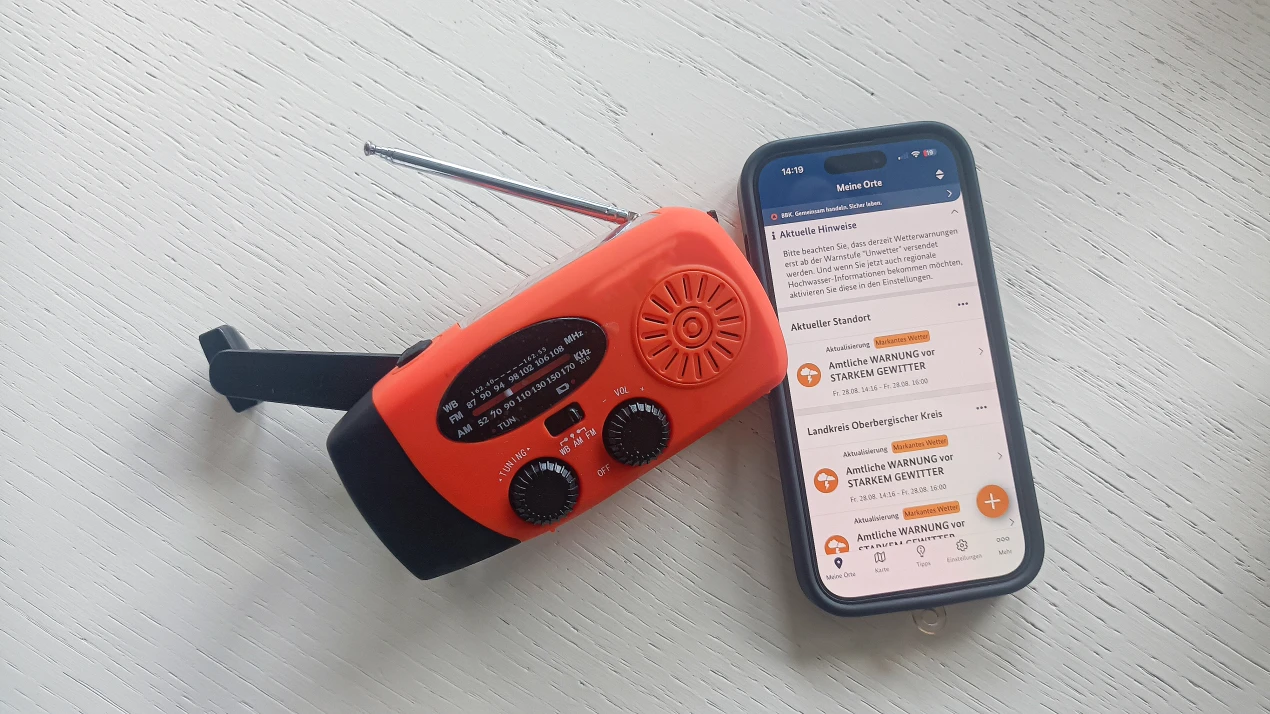 Ein orange-schwarzes Kurbelradio mit ausgezogener Antenne liegt auf einem weißen Holztisch neben einem Smartphone. Auf dem Smartphone ist eine Wetterwarn-App geöffnet, die Warnungen vor starkem Gewitter anzeigt.
