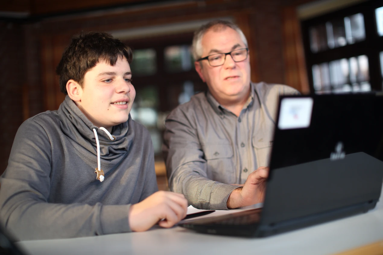Ein jüngerer und ein älterer Mann sitzen an einem Tisch und schauen gemeinsam auf einen Laptop. Der Raum im Hintergrund hat Fenster und Backsteinwände.
