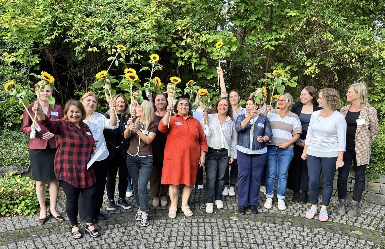 Eine Gruppe von Frauen steht auf gepflastertem Boden vor einer grünen Hecke. Sie halten Sonnenblumen in die Luft und lächeln in die Kamera.