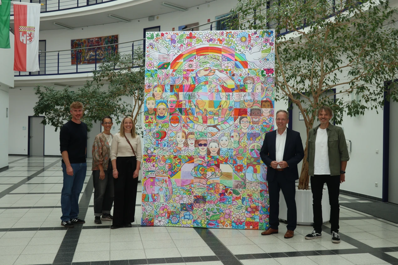 Fünf Personen stehen in einem modernen Gebäude vor einem großflächigen bunten Kunstwerk mit Motiven wie Regenbögen, Blumen, Friedenstauben und Gesichtern.