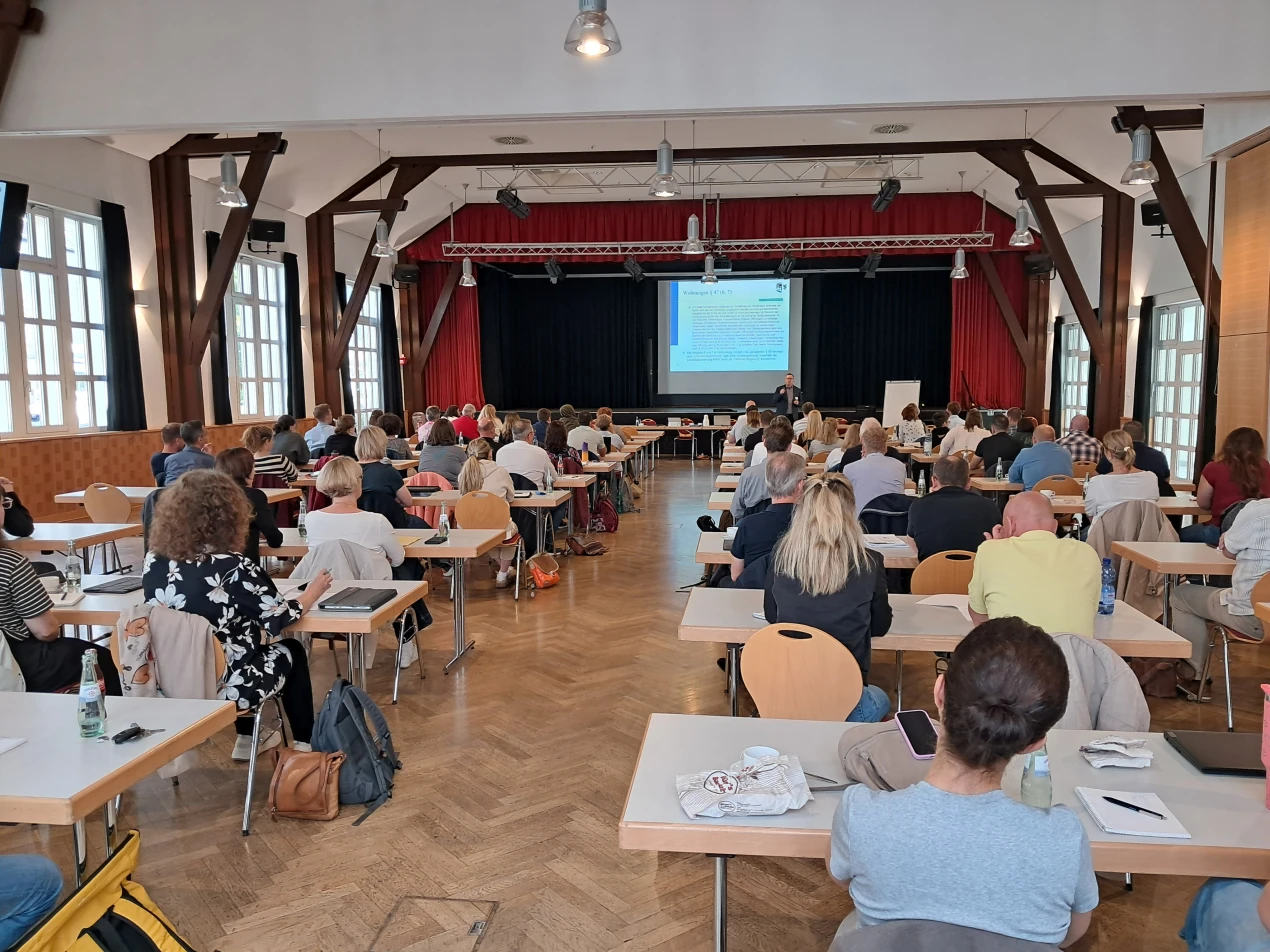 Ein großer Raum mit vielen Menschen, die an Tischen sitzen und auf eine Bühne mit einem Vortragenden und einer Präsentation blicken. Der Raum hat Holzsäulen und große Fenster.
