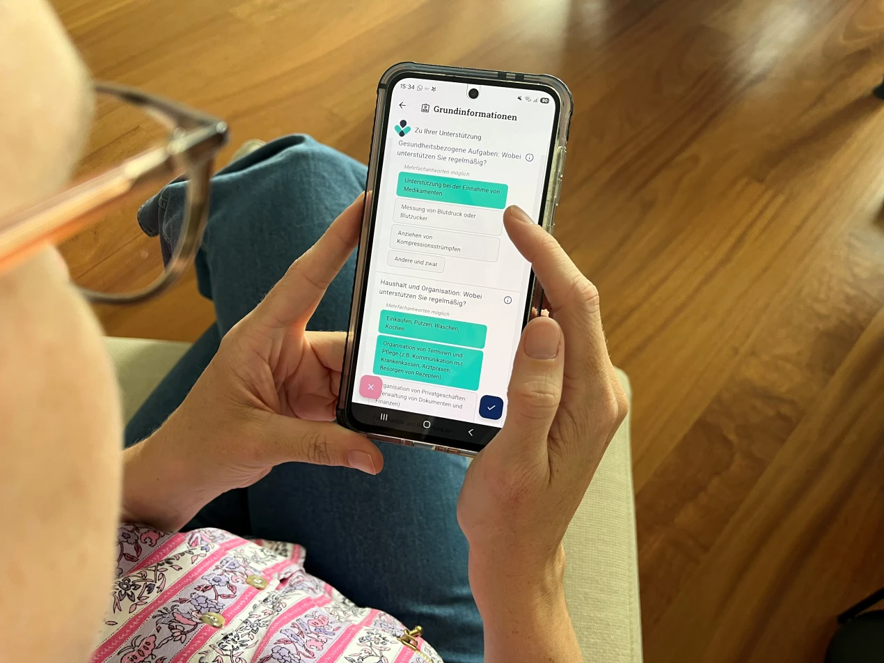 Eine Person hält ein Smartphone in der Hand und betrachtet eine App mit Formularfeldern. Die Person trägt eine Brille und ein gemustertes Hemd. Im Hintergrund ist ein Holzboden sichtbar.